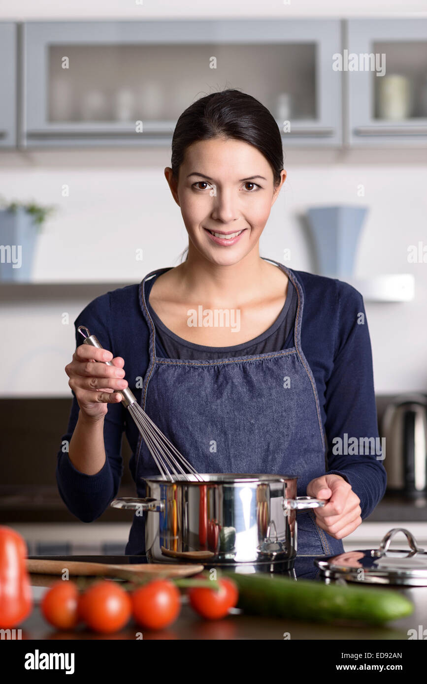Glückliche freundliche junge Hausfrau kochen Abendessen steht auf dem Kochfeld, einen Schneebesen über einem Topf halten, wie sie das Essen bereitet Stockfoto
