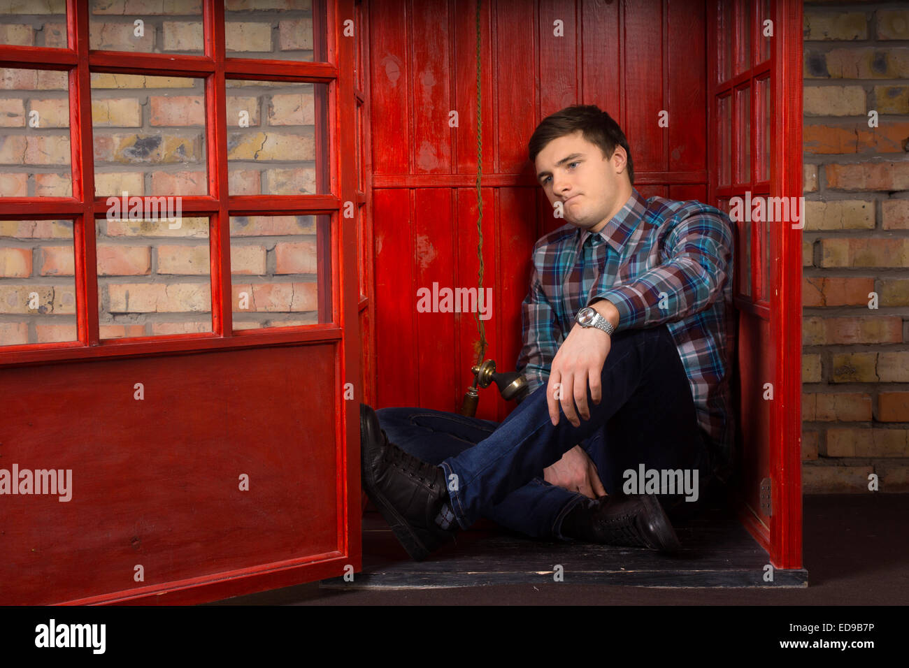 Frustrierte junge Mann einen Anruf warten, da sitzt er auf dem Boden von einer öffentlichen Telefonzelle Stirnrunzeln in Ärger Stockfoto