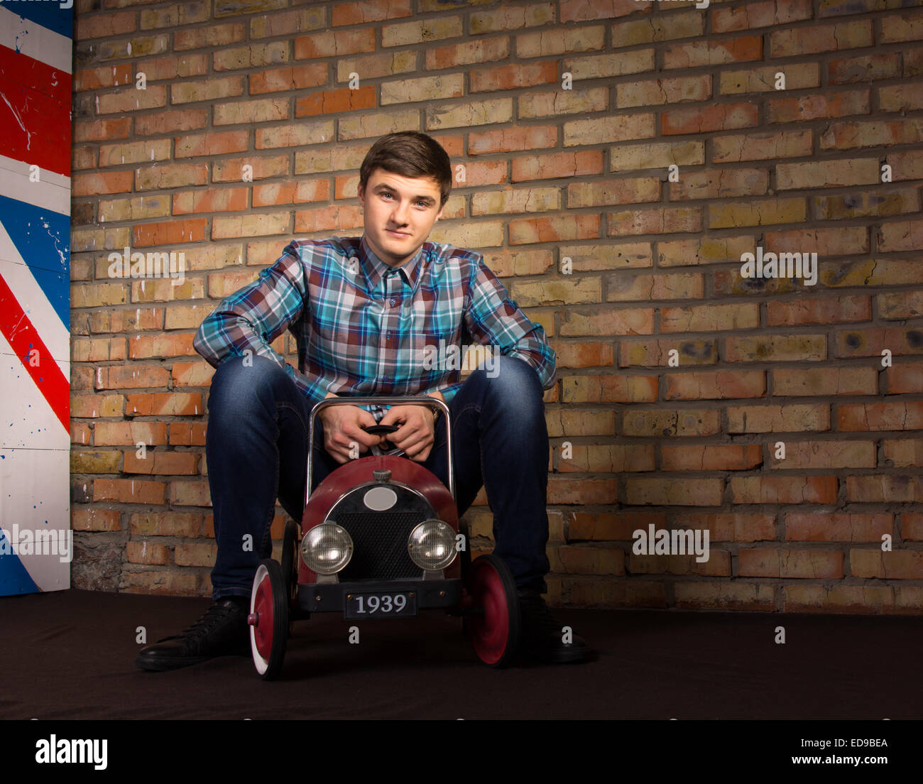 Jungen Mann in trendiges Outfit auf Spielzeug Auto sitzend und schaut in die Kamera Lächeln. Indoor mit Brick Wall Backgr erfasst Stockfoto