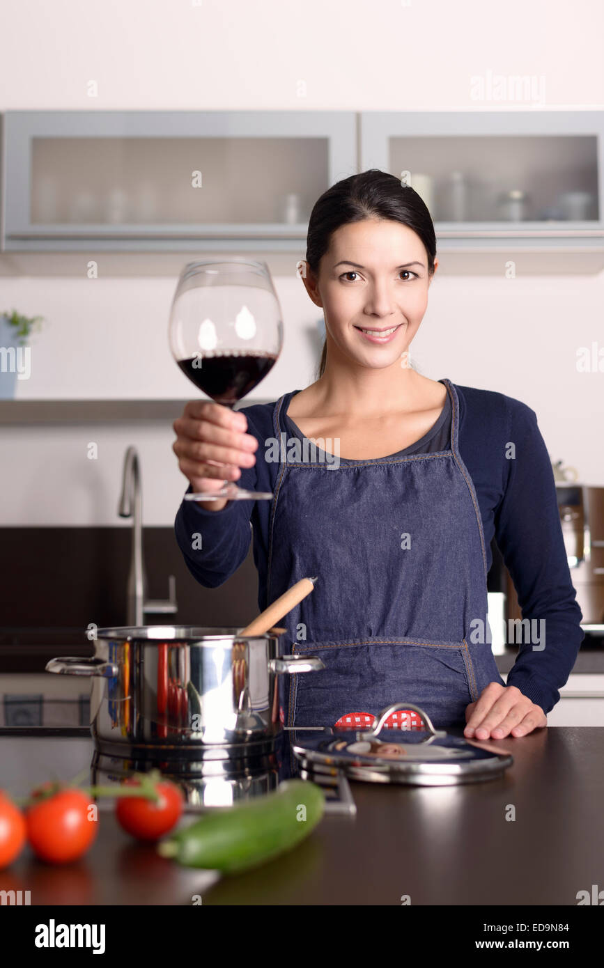 Lächelnde junge Hausfrau mit ihrem Glas Rotwein zu feiern, um die Kamera zu stoßen, während sie am Herd in der Küche steht Stockfoto