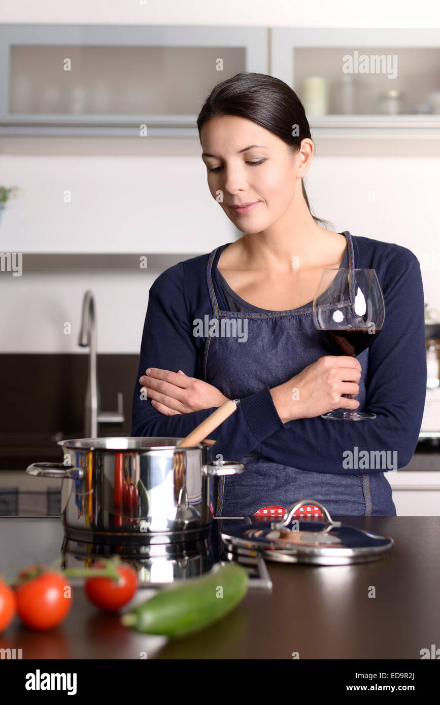 Lächelnde junge Hausfrau mit Rotwein zu feiern, während sie am Herd in der Küche vorbereiten Abendessen steht Stockfoto