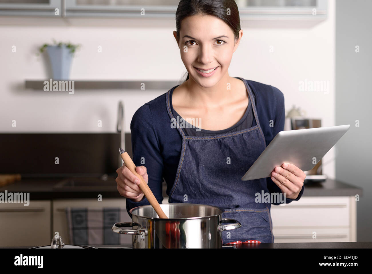 Junge Frau, die ein Rezept auf ihrem Tablet betrachten, während sie in der Küche vor einem Topf auf dem Herd steht Stockfoto