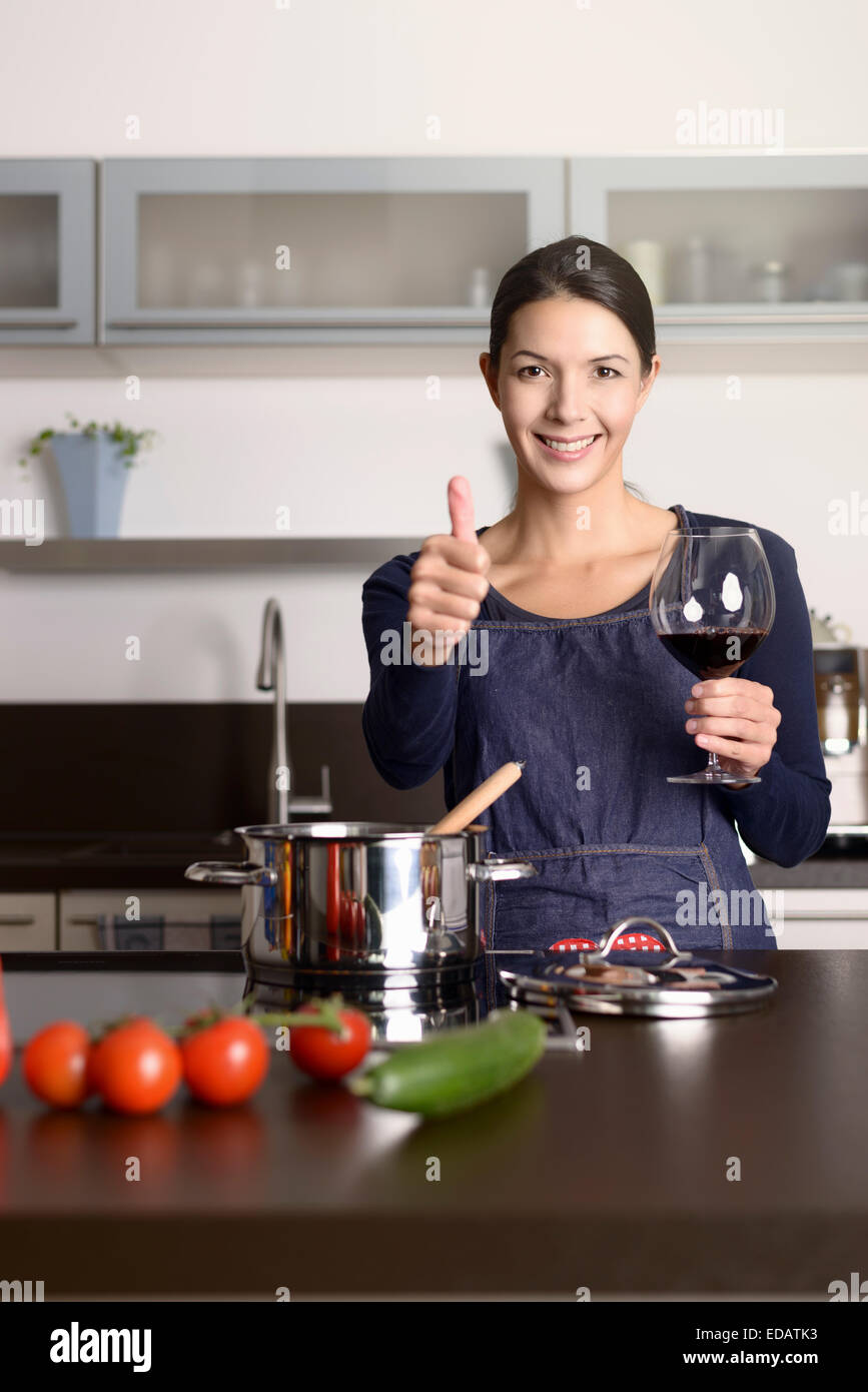 Lächelnde Hausfrau geben einen Daumen nach oben Geste, wie sie genießt ein Glas Rotwein im stehen am Herd kochen Abendessen von f Stockfoto