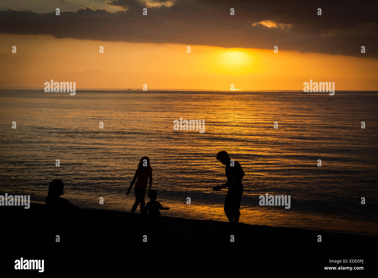 Silhouetten von Menschen, die sich vor Sonnenuntergang in Lovina, Bali, am Strand in einem bewölkten Himmel über dem Meer entspannen. Stockfoto