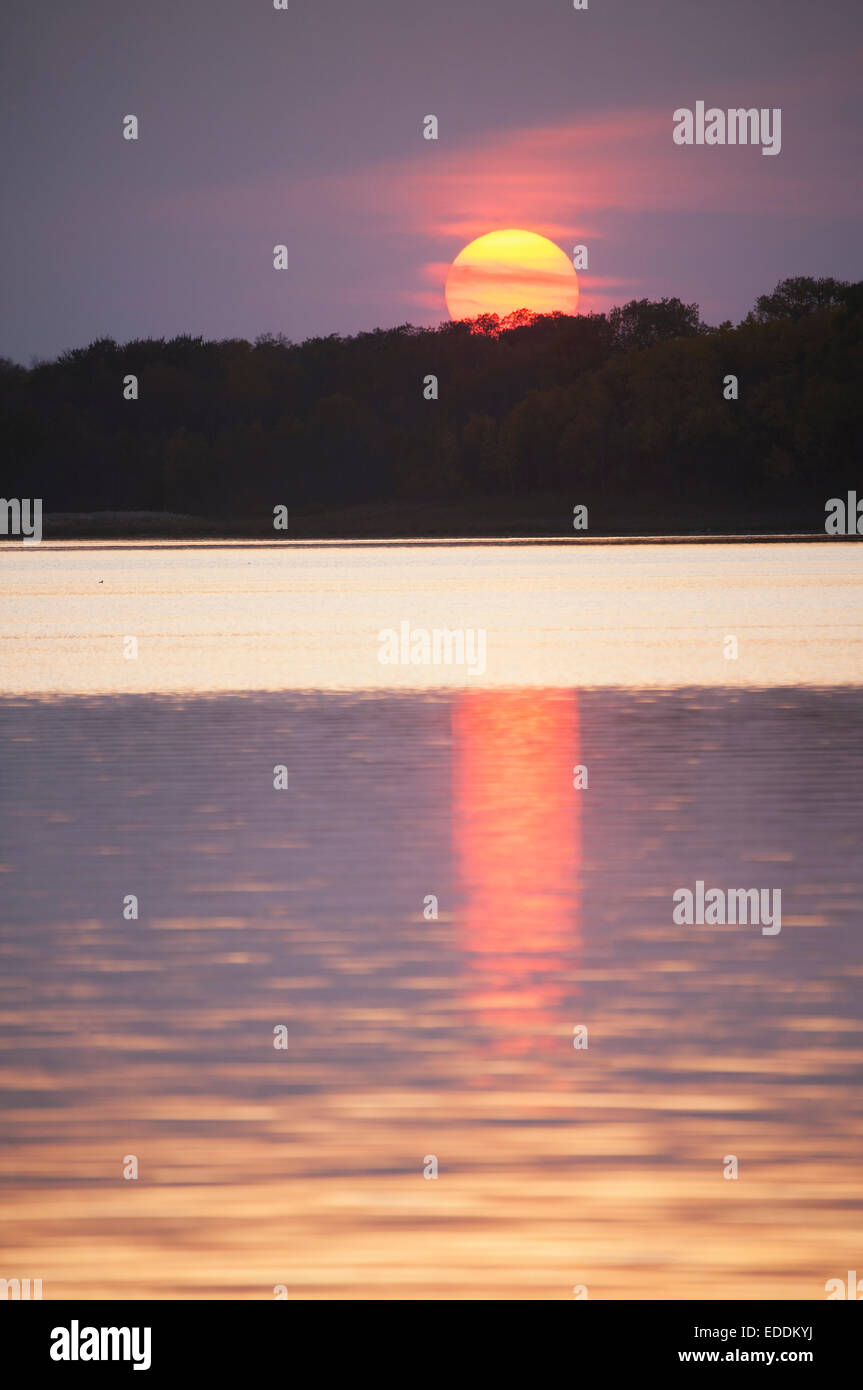 Eine orange Sonne versinkt am Horizont spiegelt sich im Wasser eines Sees. Stockfoto
