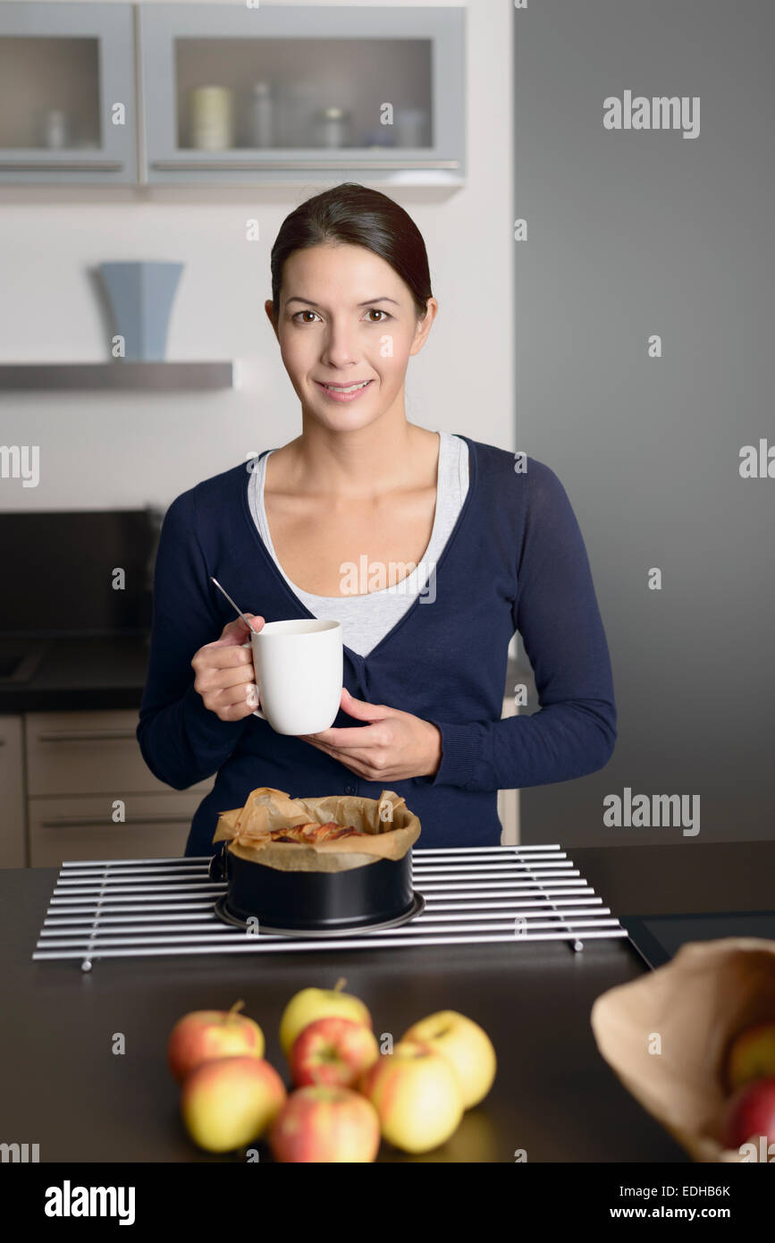 Lächelnde junge Frau Backen Apfel Torte stehen mit einer Backform, frisch aus dem Ofen nehmen, auf ein Kuchengitter entfernt, da sie f trinkt Stockfoto