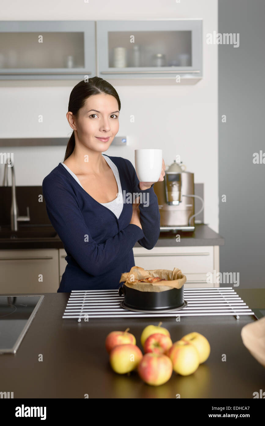 Lächelnde junge Frau Backen Apfel Torte stehen mit einer Backform, frisch aus dem Ofen nehmen, auf ein Kuchengitter entfernt, da sie trinkt Stockfoto