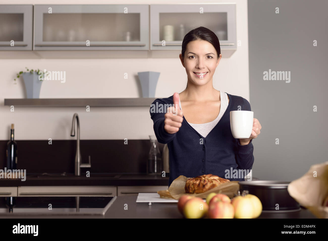 Glücklich attraktive junge Köchin geben einen Daumen nach oben und Erfolg wie sie in ihrer Küche steht, Kaffee zu trinken Stockfoto