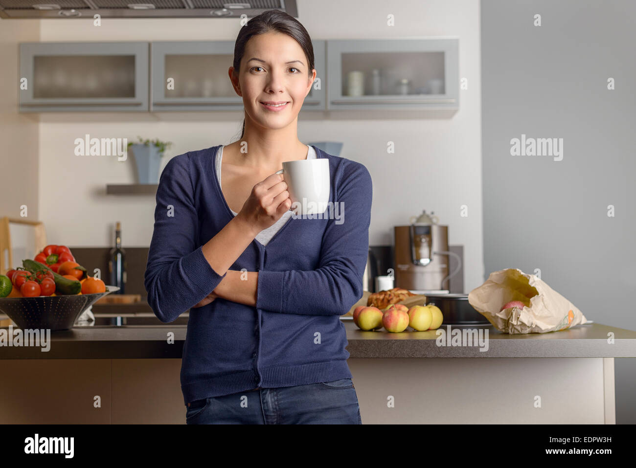 Glückliche zufriedene Hausfrau in ihrer Küche geben der Kamera ein warmen freundliches Lächeln, wie sie mit einer Tasse Kaffee mit frischen entspannt Stockfoto