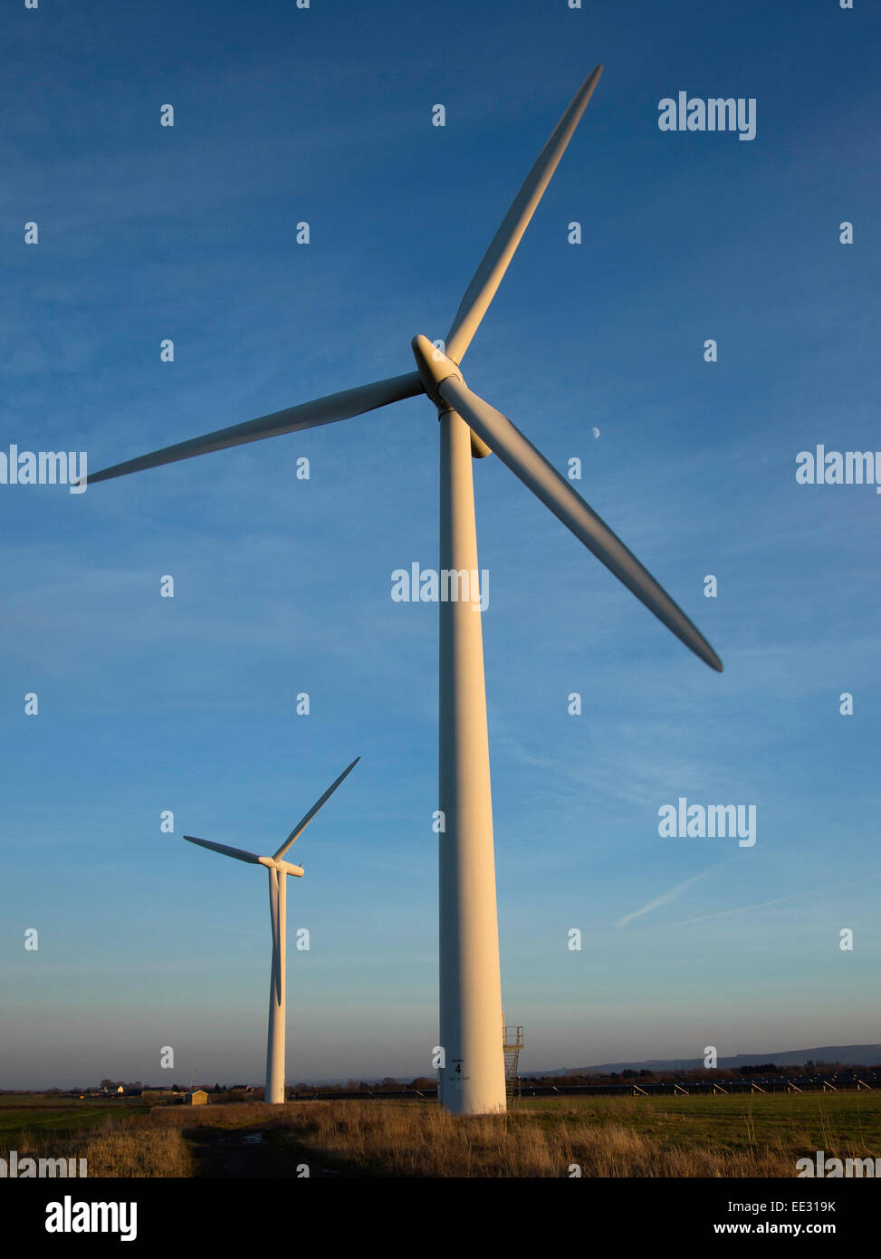 Zwei Windkraftanlagen, gegen einen blauen Himmel und der Mond an einem Wintertag in der Westmill Windfarm kooperative in Oxfordshire, Vereinigtes Königreich. Stockfoto