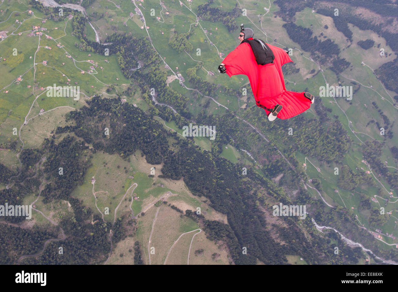 Eine Wingsuit Pilot in seinem roten Vogelmann Anzug fliegt über grüne Felder. So verdoppelt er seine Flugzeit und er lächelt. Stockfoto