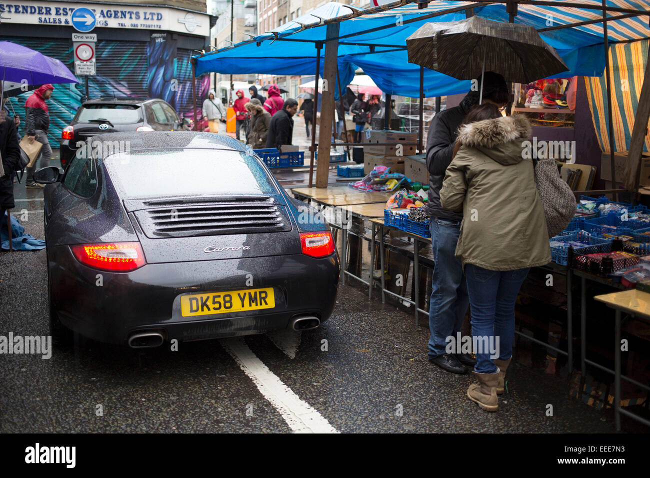 Regnerischen Tag auf Brick Lane im East End von London, UK. Porche 911 fährt durch den Markt, wie Menschen über den Tag zu gehen. Es ist eine Illustration der Besitzenden und Habenichtse wie ein Sportwagen Zehntausende von Pfund Wert vorbeifährt Stände super billig verkaufen. Stockfoto