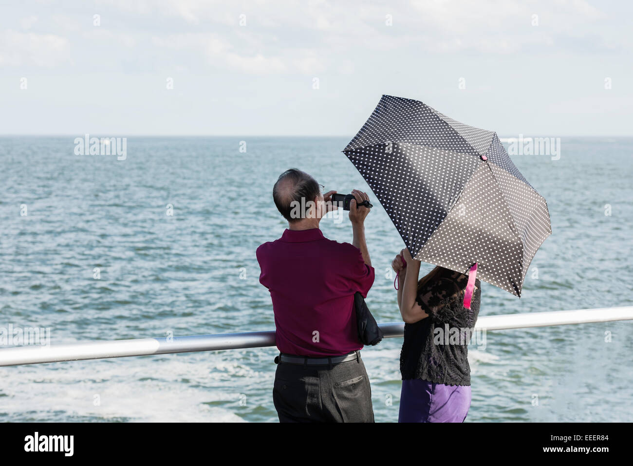 Rückansicht des älteren asiatischen Mann mit Camcorder und Frau unter einem Polk versteckte-a-dot Dach stehend an einem Geländer an den Atlantischen Ozean. Stockfoto