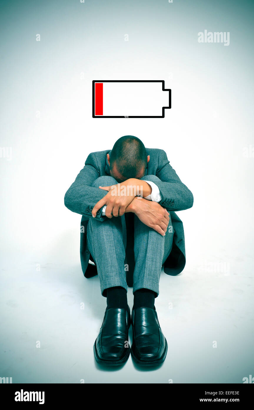 ein junger Geschäftsmann zusammengerollt in den Boden mit dem Kopf zwischen seine Knie und ein Beispiel für eine schwache Batterie Stockfoto