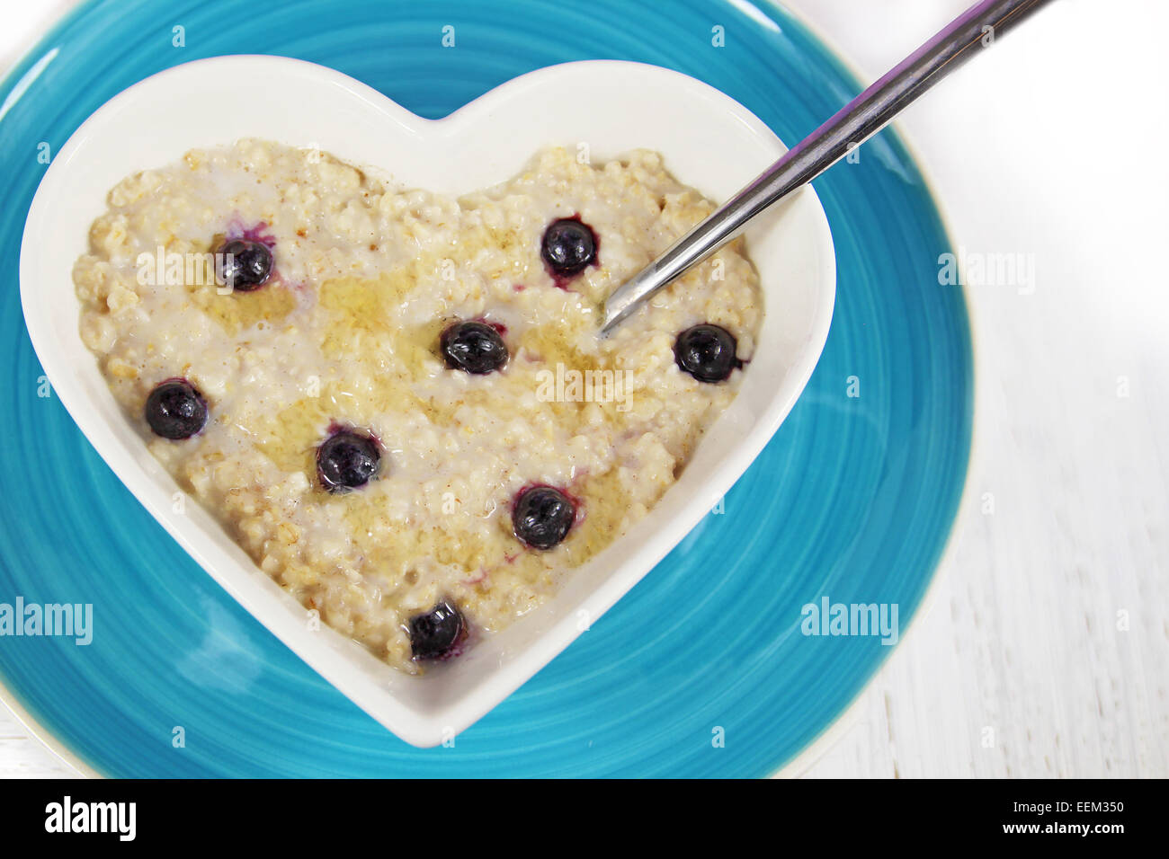 Heidelbeer-Porridge oder Haferflocken mit Honig in eine herzförmige Schüssel mit einem Löffel. Ruht auf der blauen Platte auf einem weißen Hintergrund rustikal. Stockfoto