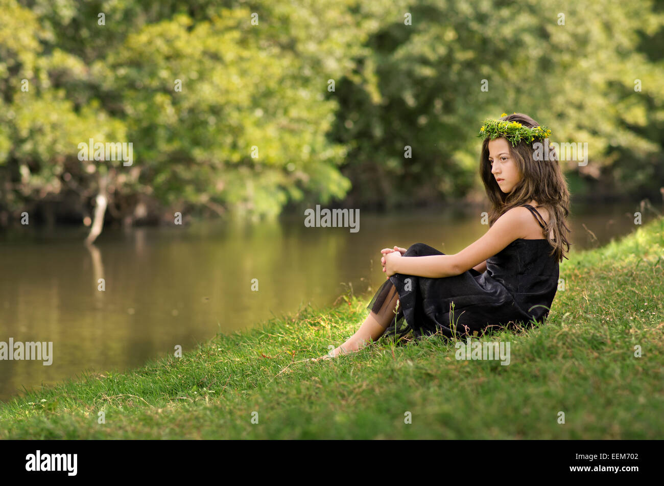 Niedliche kleine Mädchen (10-11) mit Kranz entspannend am Flussufer Stockfoto