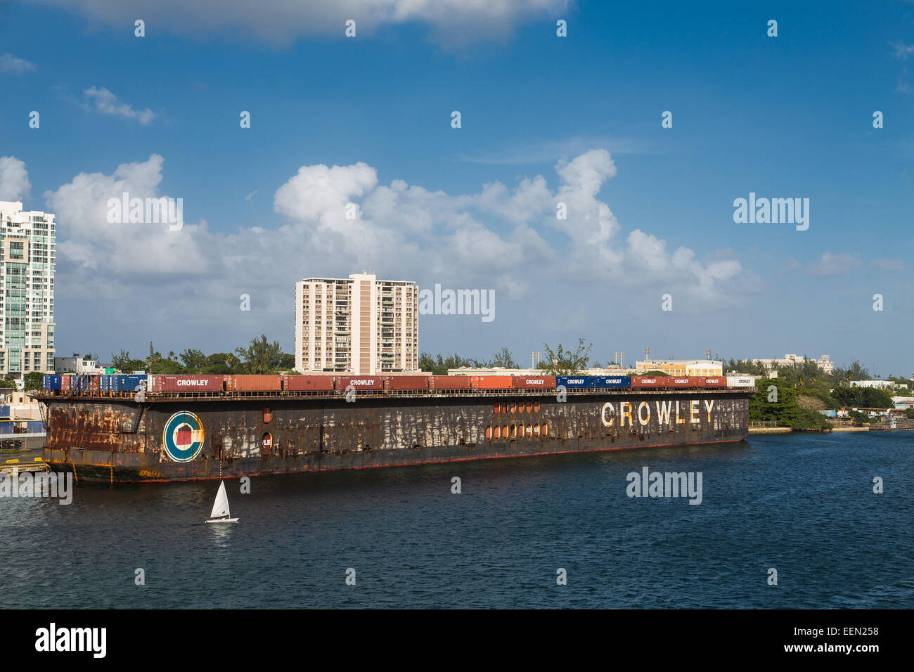 Ein kleines weißes Segelboot im blauen Wasser vor der Küste von San Juan von einem riesigen rostigen Frachter Stockfoto