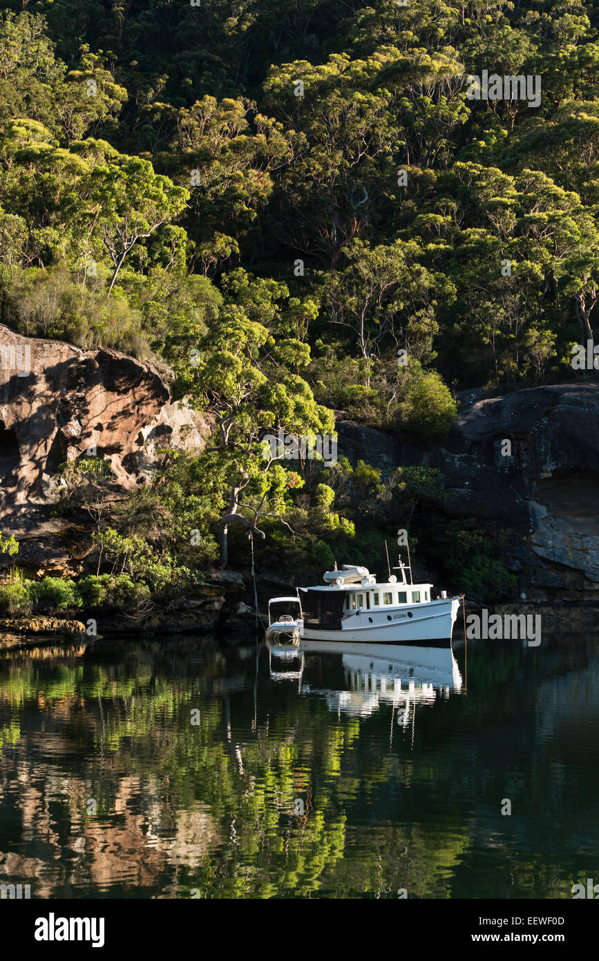 Ein Motorboot am Anker und am frühen Morgensonnenlicht an bewaldeten Hängen spiegeln sich in den ruhigen Gewässern des Smiths Creek, Broken Bay. Stockfoto