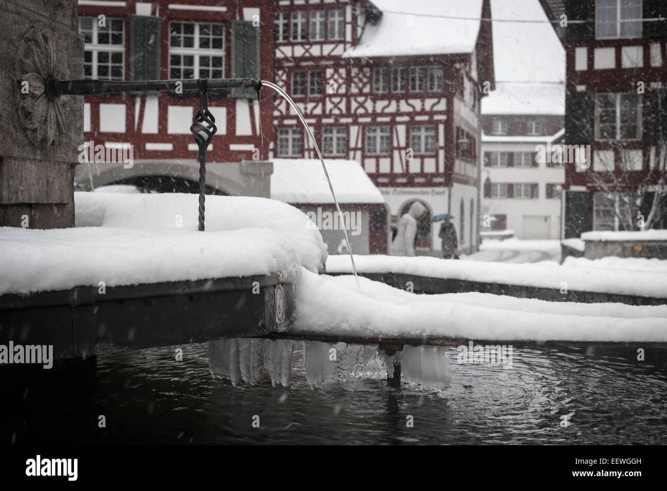 Brunnen mit Schnee und Eiszapfen mit typisch Schweizer Bar befindet sich im Hintergrund, Bülach, Schweiz. Stockfoto