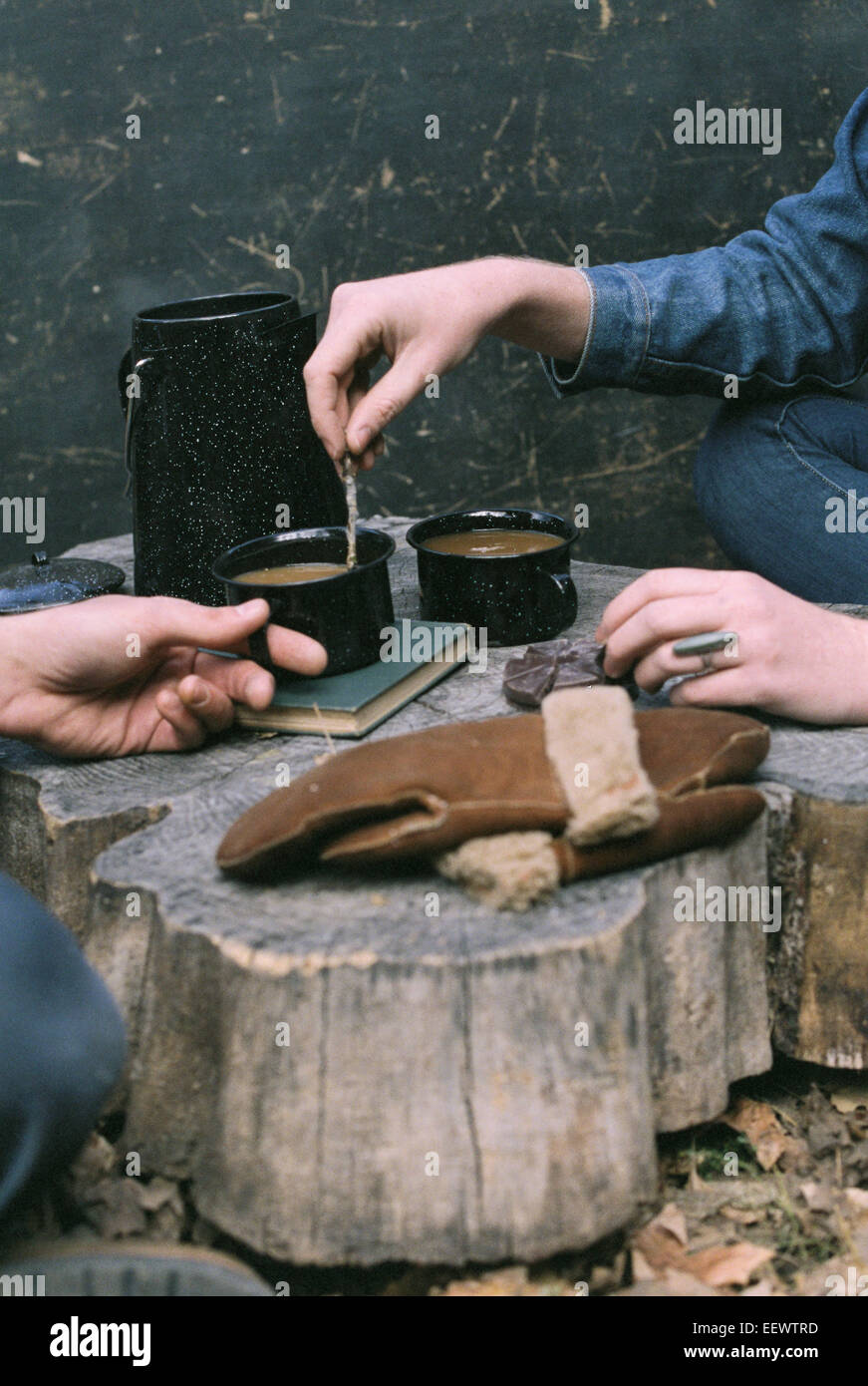 Paar beim Kaffee in einem Wald, zwei Tassen Kaffee, eine Kaffeemaschine und ein paar Handschuhe auf einem Baumstamm. Stockfoto