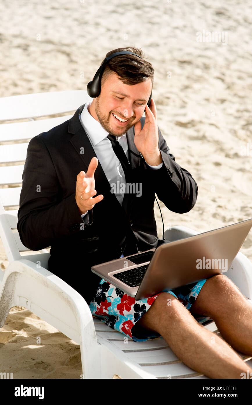 Geschäftsmann, gekleidet in Anzug und Shorts mit Videoanruf mit Laptop auf der Liege am Strand Stockfoto