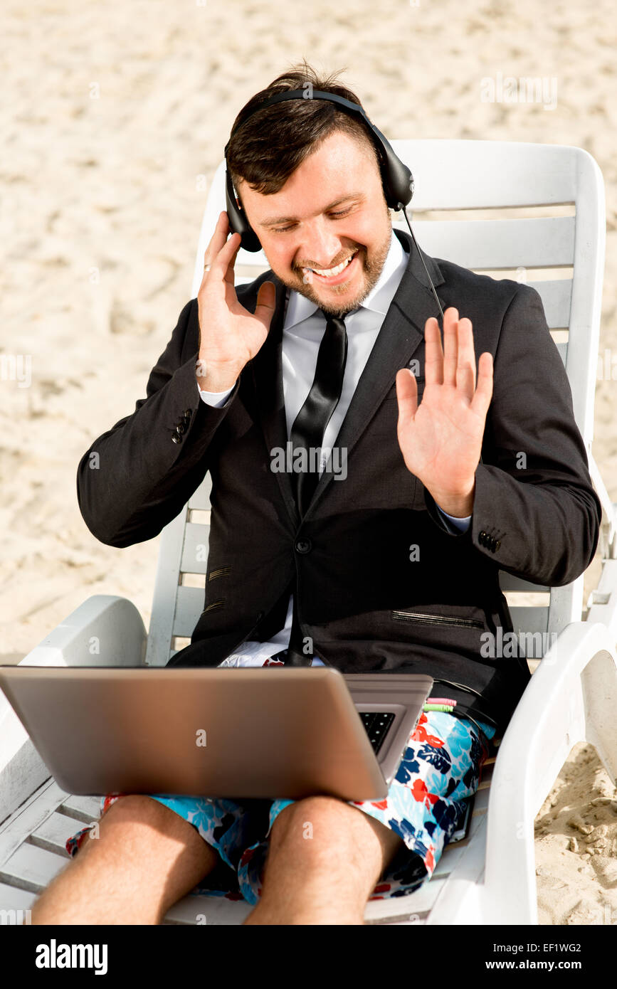 Geschäftsmann, gekleidet in Anzug und Shorts mit Videoanruf mit Laptop auf der Liege am Strand Stockfoto