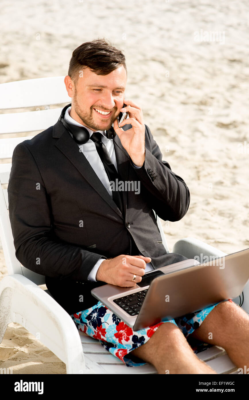 Geschäftsmann, gekleidet in Anzug und Shorts arbeiten mit Laptop auf dem Liegestuhl am Strand Stockfoto