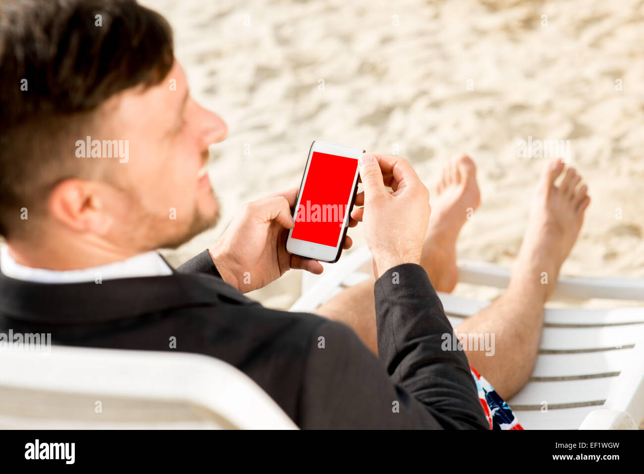 Geschäftsmann hält Handy mit leeren Bildschirm sitzen auf der Sonnenbank gekleidet in Anzug und Shorts am Strand Stockfoto