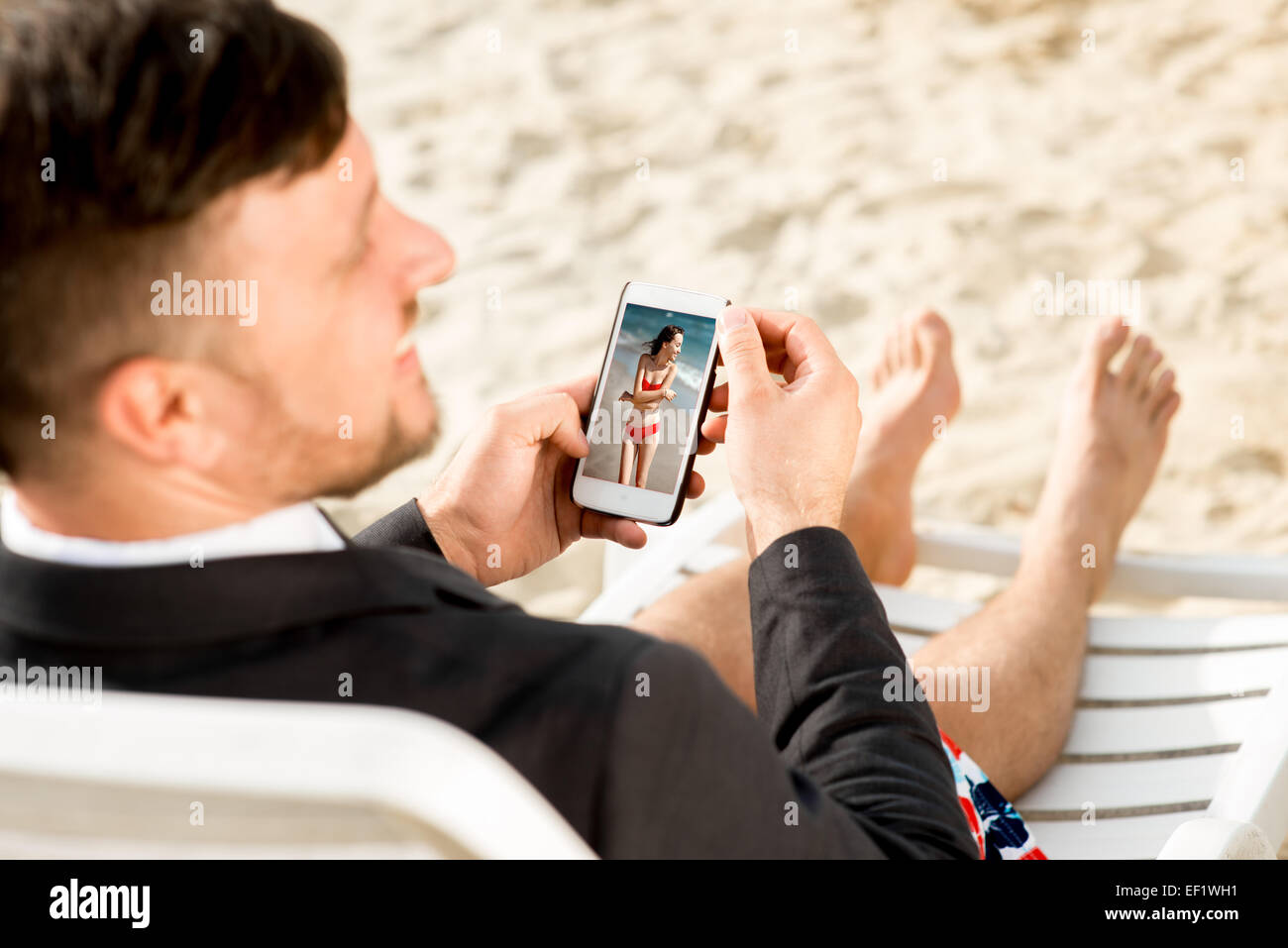 Geschäftsmann hält Handy mit Foto der Freundin auf der Liege sitzen gekleidet in Anzug und Shorts am Strand Stockfoto