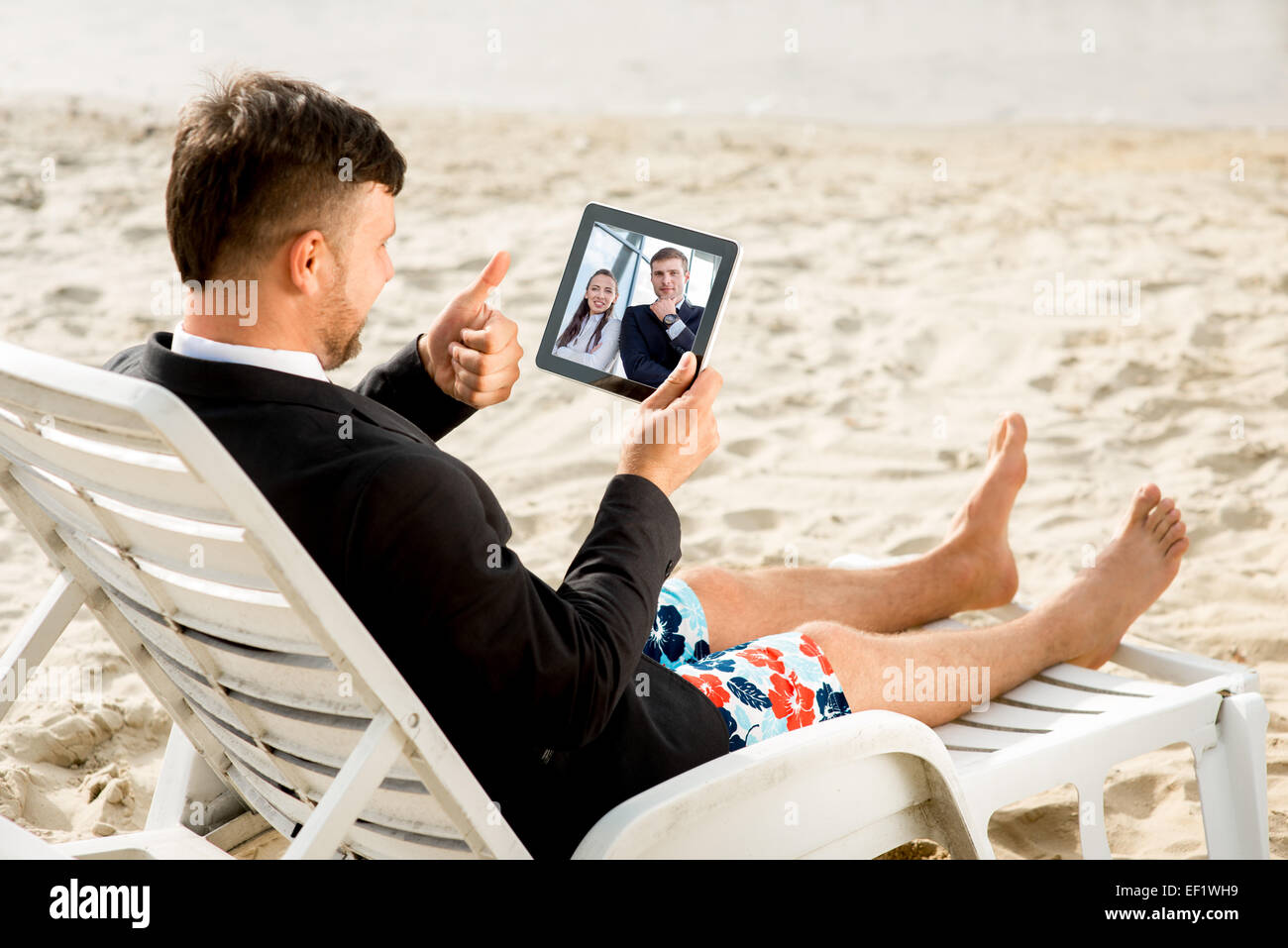 Geschäftsmann, gekleidet in Anzug und Shorts mit Videoanruf mit digital-Tablette auf der Liege am Strand Stockfoto