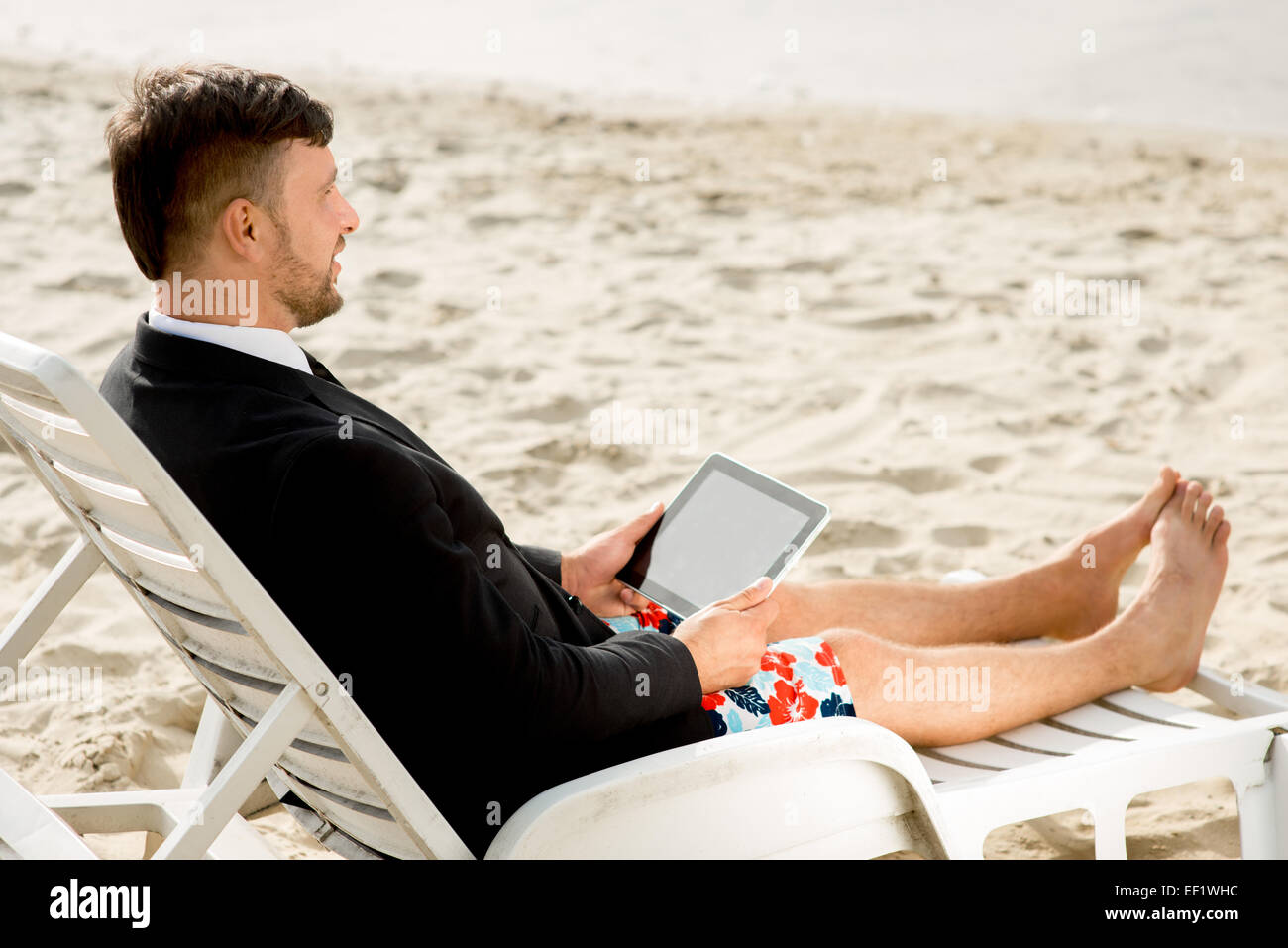 Geschäftsmann, gekleidet in Anzug und Shorts arbeiten mit Laptop auf dem Liegestuhl am Strand Stockfoto