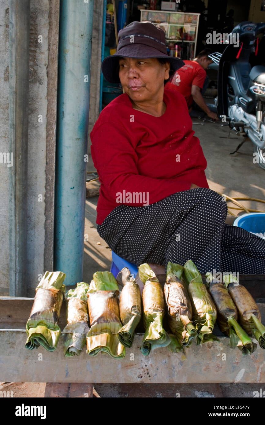 Eine Frau Straßenhändler verkauft Klebreis in Pandan Blätter eingewickelt & gegrillt auf Kohlenfeuer auf einer Stadtstraße in Kampong Cha Stockfoto