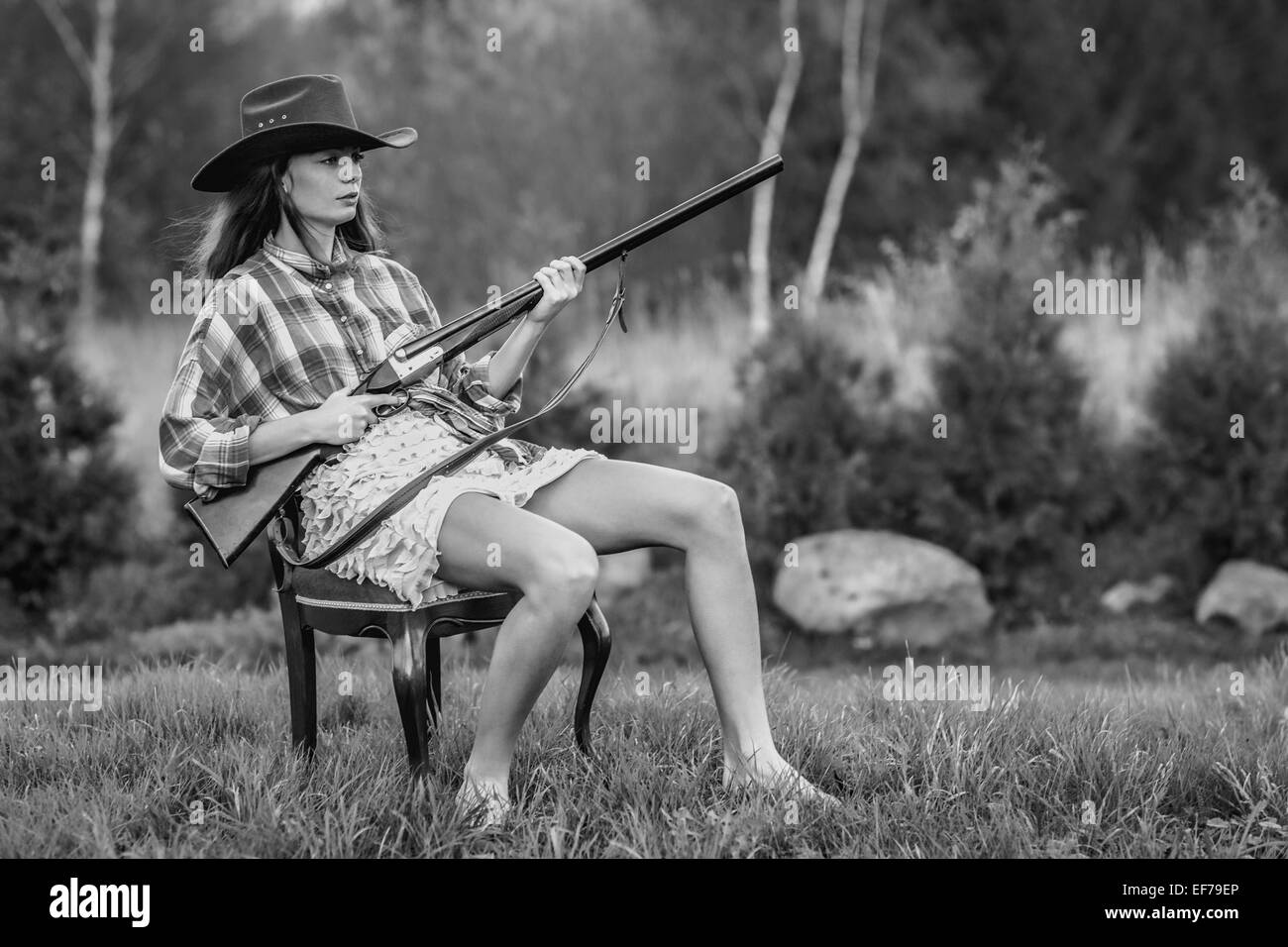 Schwarz / weiß Fotografie eines Mädchens in Cowboy-Hut mit einer Schrotflinte in einem Hinterhof in einem ausgefallenen Sessel sitzend. Stockfoto