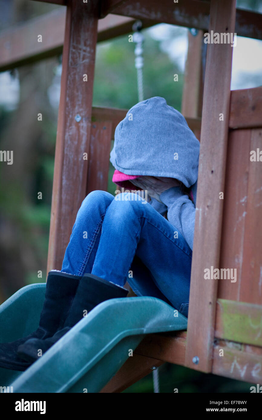 Trauriges Kind in Hoodie mit Gesicht versteckt in Händen, sitzt alleine auf einem Spielplatz in Verzweiflung. Stockfoto