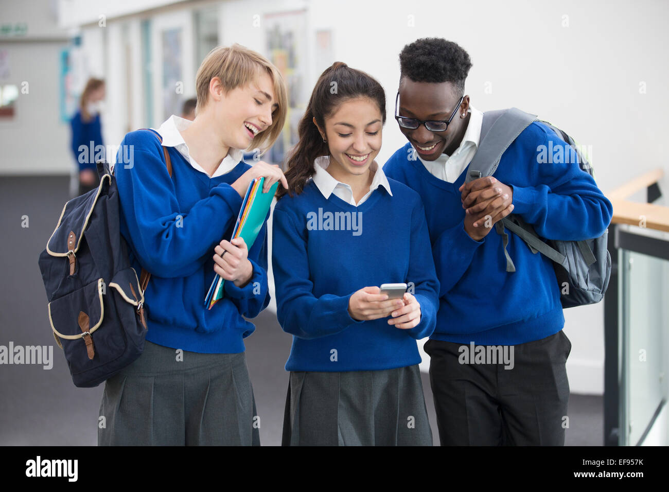 Drei lachende Studenten tragen blaue Uniformen mit Handy in Schule Flur Stockfoto