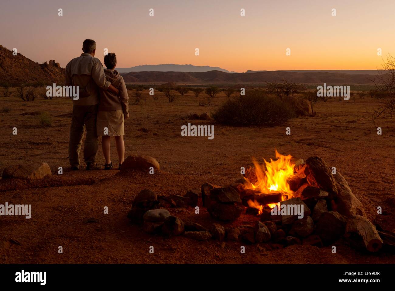 Eine romantische Aufnahme von ein paar genießt die untergehende Sonne am Mowani Mountain Camp, Damaraland, Namibia, neben ihrem Lagerfeuer. Stockfoto