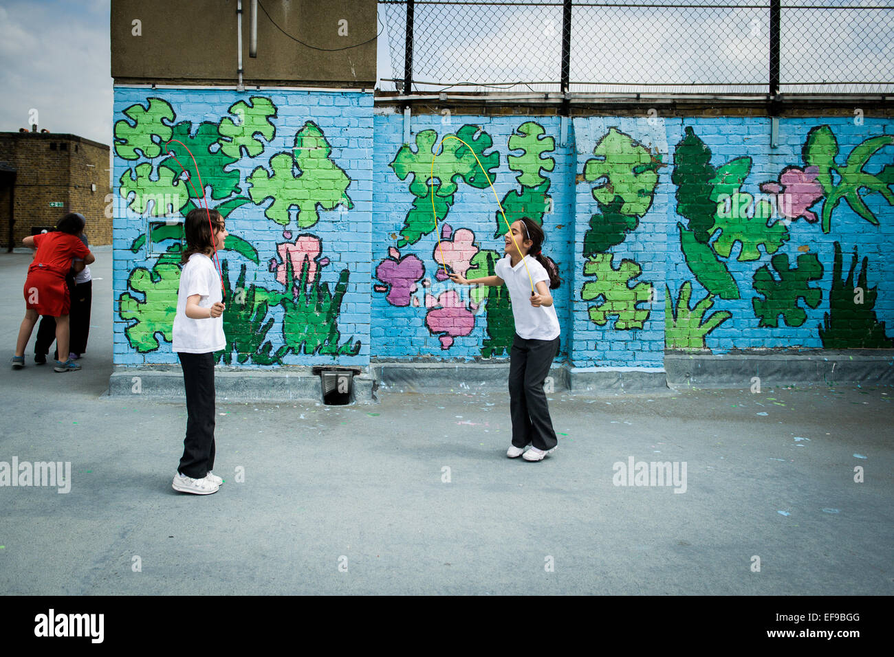 2 school Mädchen überspringen in den Spielplatz auf dem Dach an Ihre innere Stadt Grundschule mit Graffiti an der Wand hinter Stockfoto
