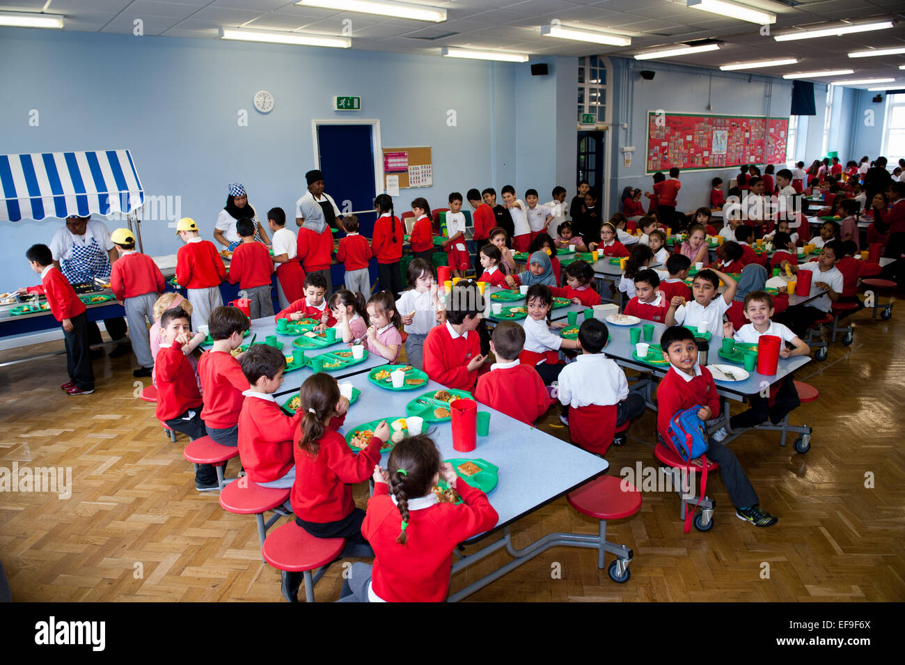 Schulabendessen - Mittagszeit in der britischen State Primary School Lehrer und Grundschulkinder in der Primary School Stockfoto