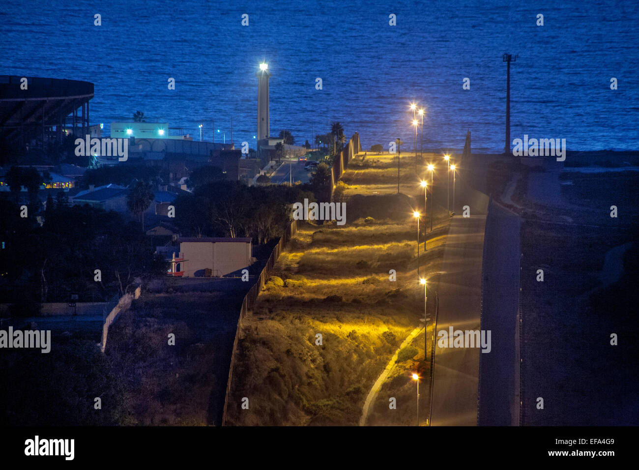 Natrium Leuchten Dampf die hügelige Staatsgrenze zwischen den USA und in Tijuana, Mexiko auf der linken Seite. Hinweis: Pazifischer Ozean und Leuchtturm im Hintergrund. Stockfoto