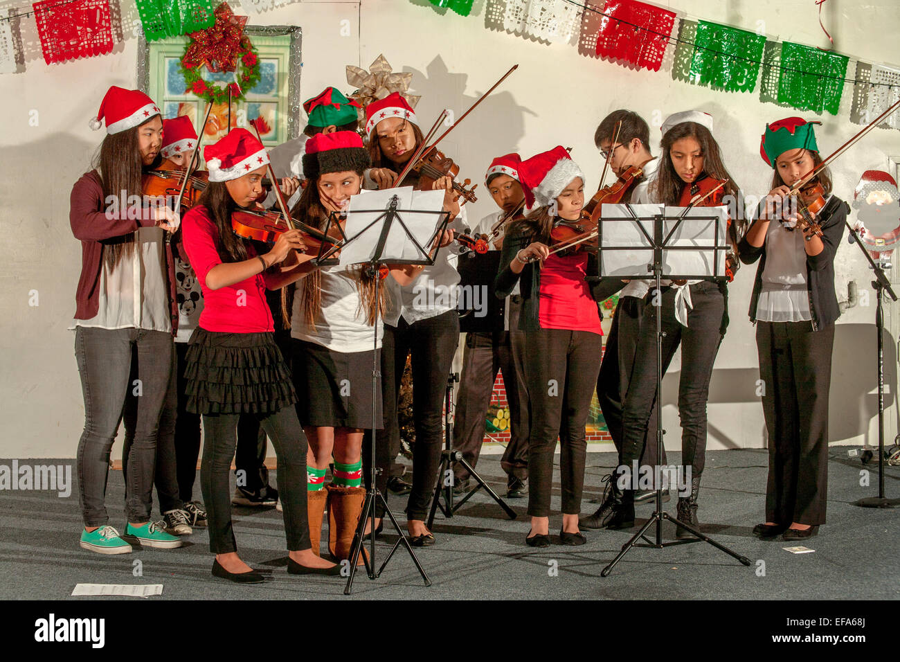 Ausgestattet in Santa Hüte, spielen junge Hispanic Musikstudenten Geigen an einem Weihnachtskonzert in einem Santa Ana, CA, "Barrio" Gemeindezentrum Auditorium. Hinweis: freiwillige asiatisch-amerikanische High School Musik Mentoren und mexikanischen Nationalfarben. Stockfoto