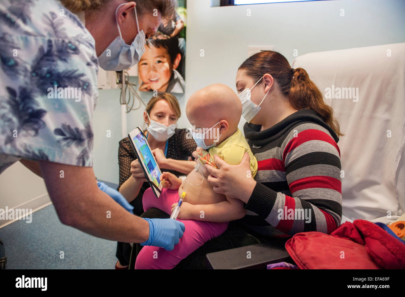 Ein Kind Leben Specialist bei CHOC Kinderkrankenhaus in Orange, CA, hält ein Computerprogramm Tablet um einen jungen hispanischen Mädchen Patienten eine Port-Zugriffsverfahren für Chemotherapie abzulenken. Hinweis: Masken, Krankenschwester und Glatze. Stockfoto