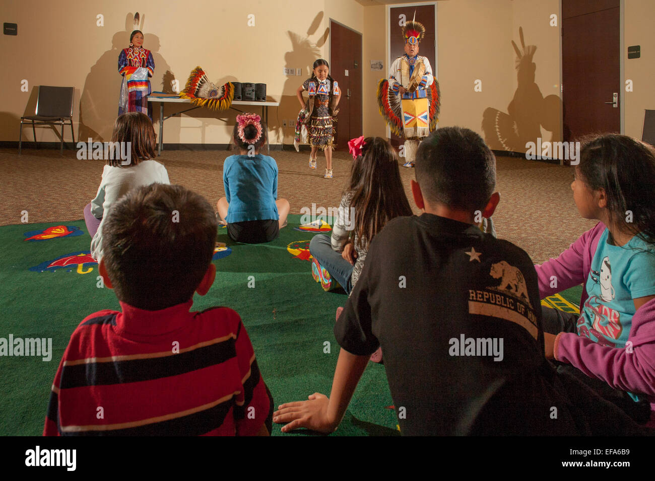 Wie Kinder im Publikum zu sehen, führt eine 7-Year-Old Navajo indische Tänzerin in vollen tribal Kostüm Jingle Dress Dance an einem Abend der Kultur der amerikanischen Ureinwohner in der Laguna Niguel, CA, Stadtbibliothek. Hinweis: kostümierte Vater und Schwester. Stockfoto
