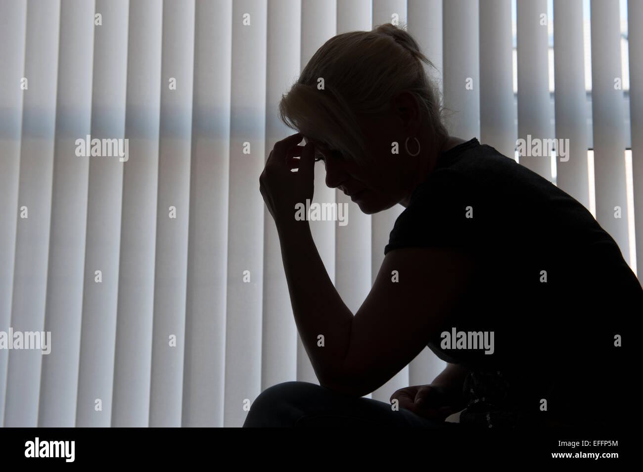 Eine Frau mittleren Alters Silhouette gegen Fenstervorhänge lehnt sich über den Kopf in ihre Hände. Stockfoto