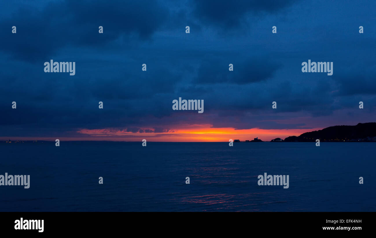 Ein goldener Sonnenaufgang wirft den Leuchtturm und Rettungsboot wirft an Mumbles scharfe Konturen unter einem stürmischen Winterhimmel. Stockfoto