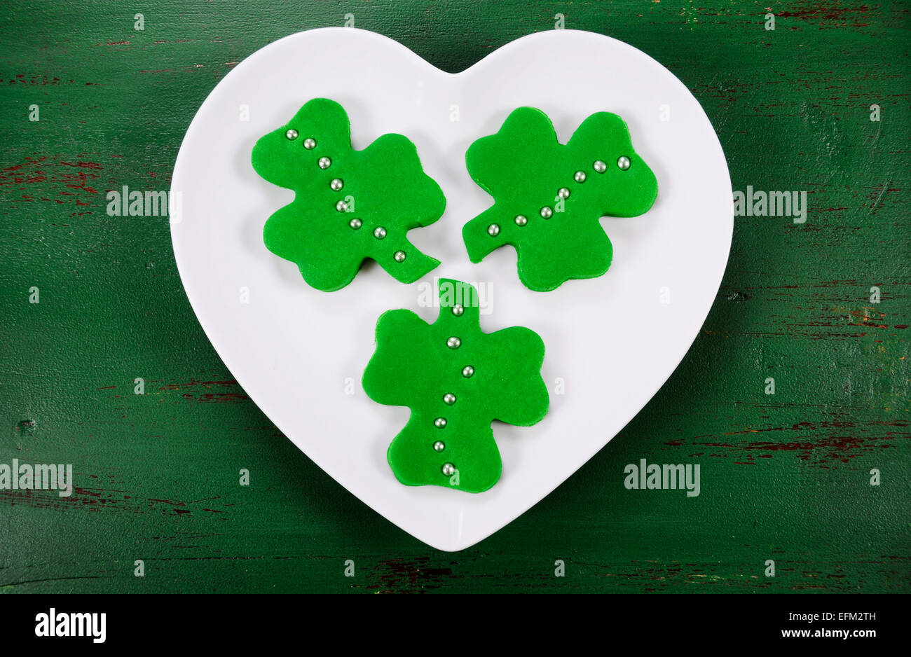Happy St Patricks Day Kleeblatt gestalten grüne Fondant Cookies auf weißes Herz Form Platte auf Vintage-Stil grün Holztisch. Stockfoto