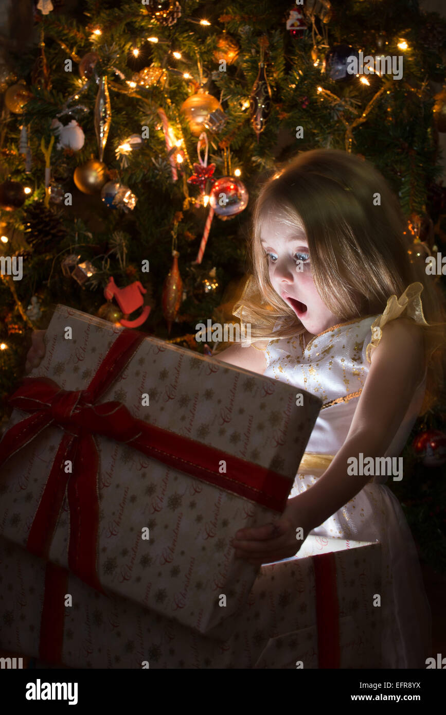 Eine junge, aufgeregte, kaukasische Mädchen ist aufgeregt und überrascht beim Öffnen von einer Weihnachts-Box durch ein traditionell geschmückten Weihnachtsbaum Stockfoto