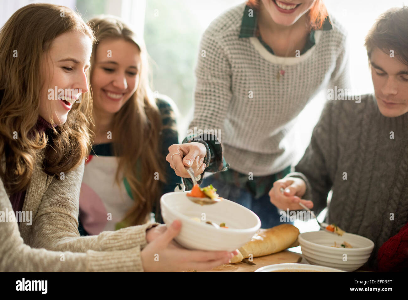 Vier Personen sitzen und stehen an einem Tisch, eine Frau mit Essen in eine Schüssel geben. Stockfoto