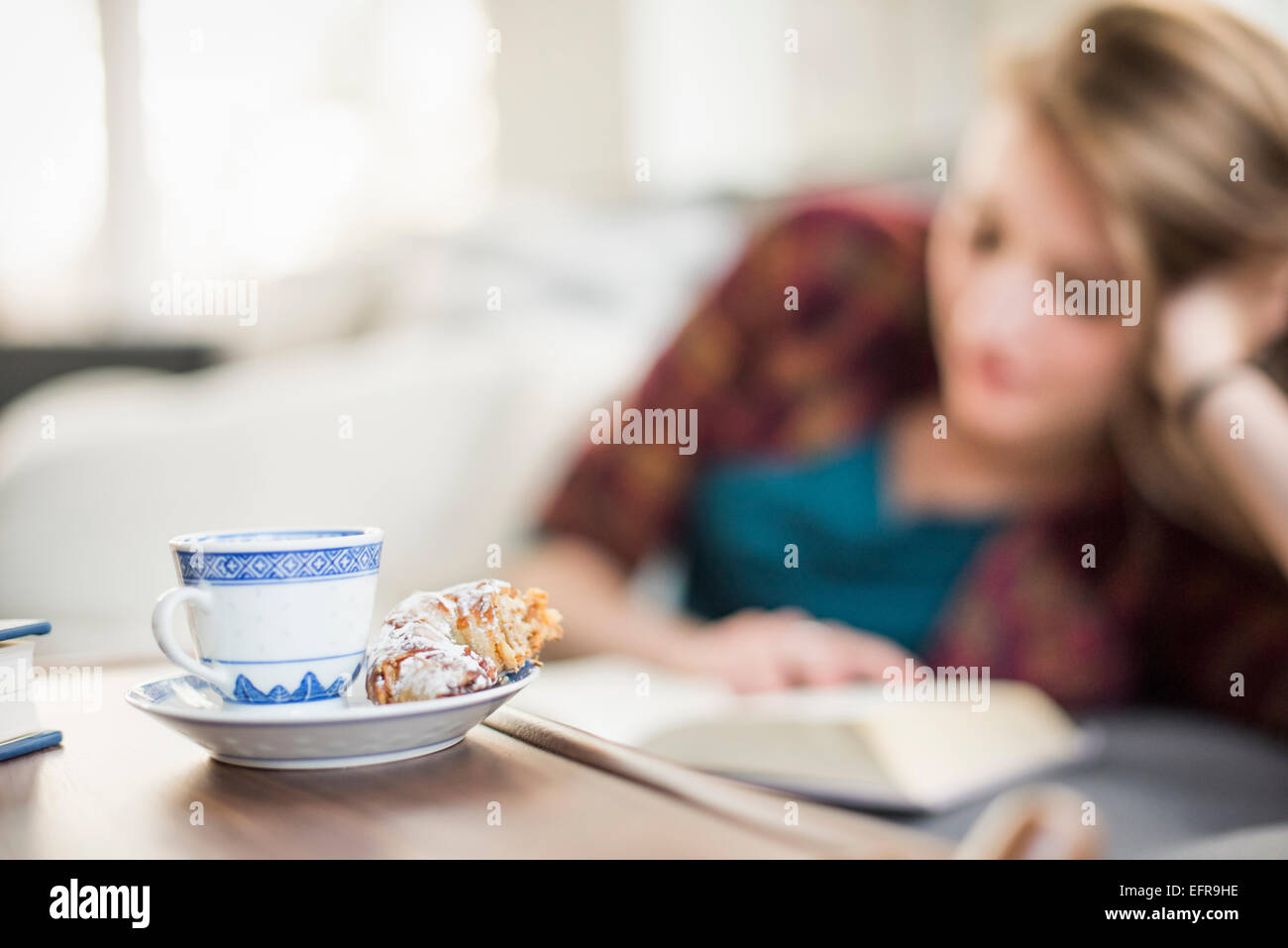 Junge Frau sitzt auf einem Sofa, ein Buch zu lesen. Eine Tasse und Untertasse mit einem Croissant auf einem Tisch. Stockfoto