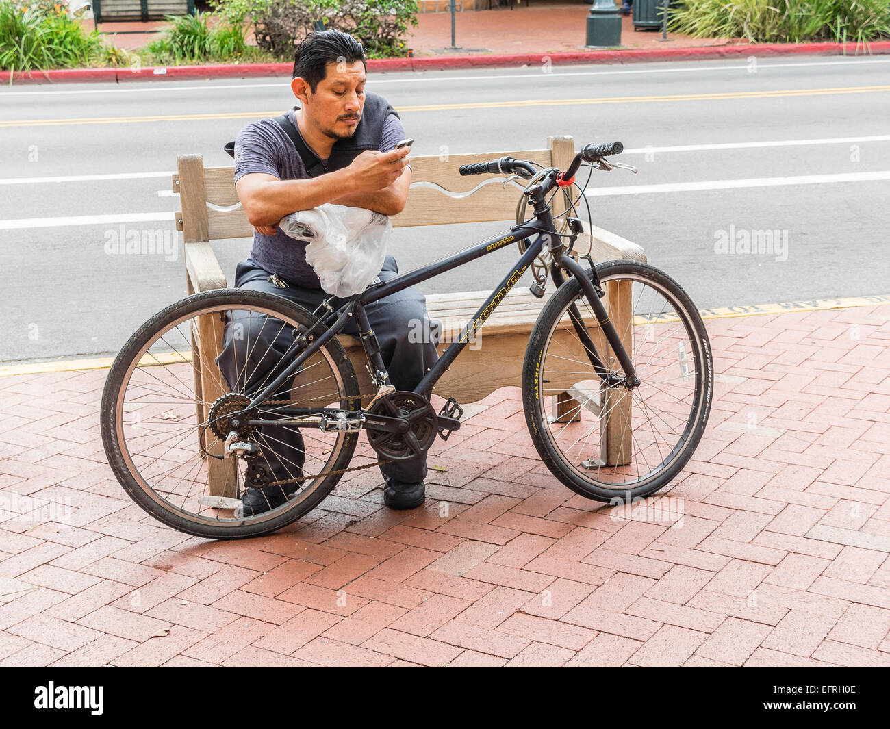 Ein mittleren Alters Hispanic Mann sitzt auf einer Bank der Stadt auf einem Bürgersteig neben State Street Überprüfung sein Handy mit seinem Fahrrad in Stockfoto