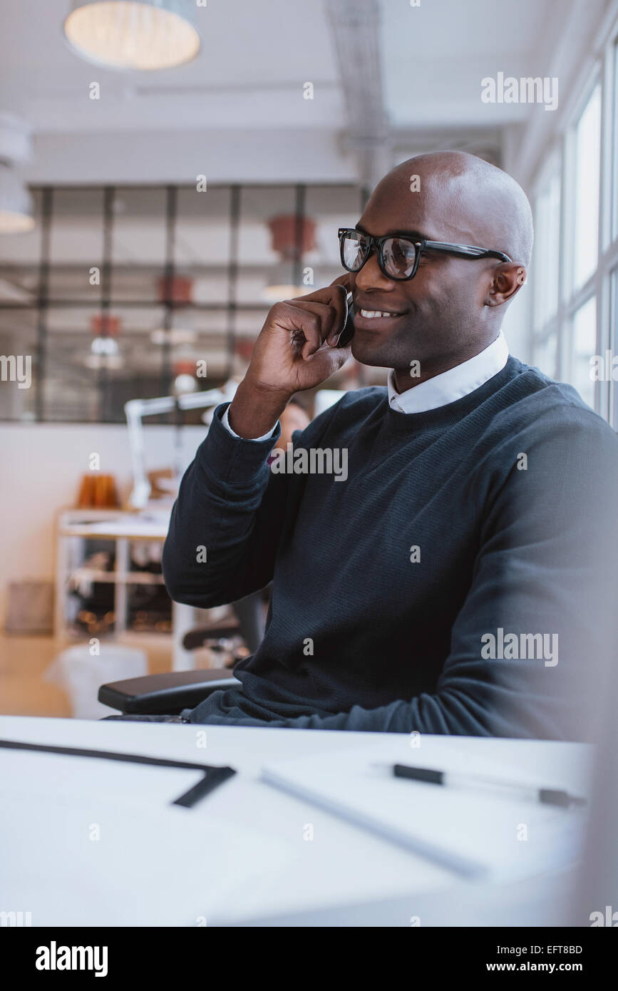 Glücklicher junge afrikanische Mann sitzt an seinem Schreibtisch auf seinem Handy im Büro sprechen. Afrikanische Executive mit Handy. Stockfoto